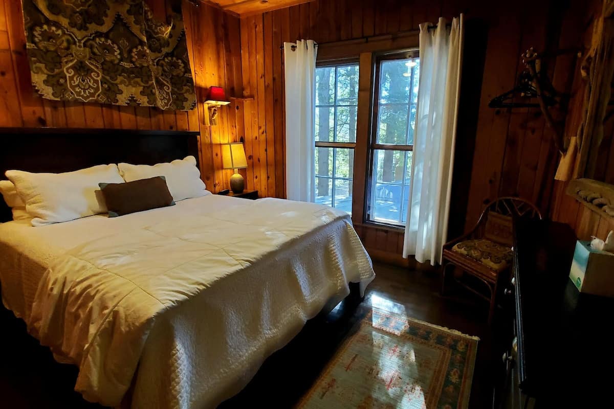 A wooden room with a bed, a chair, and a window with curtains.