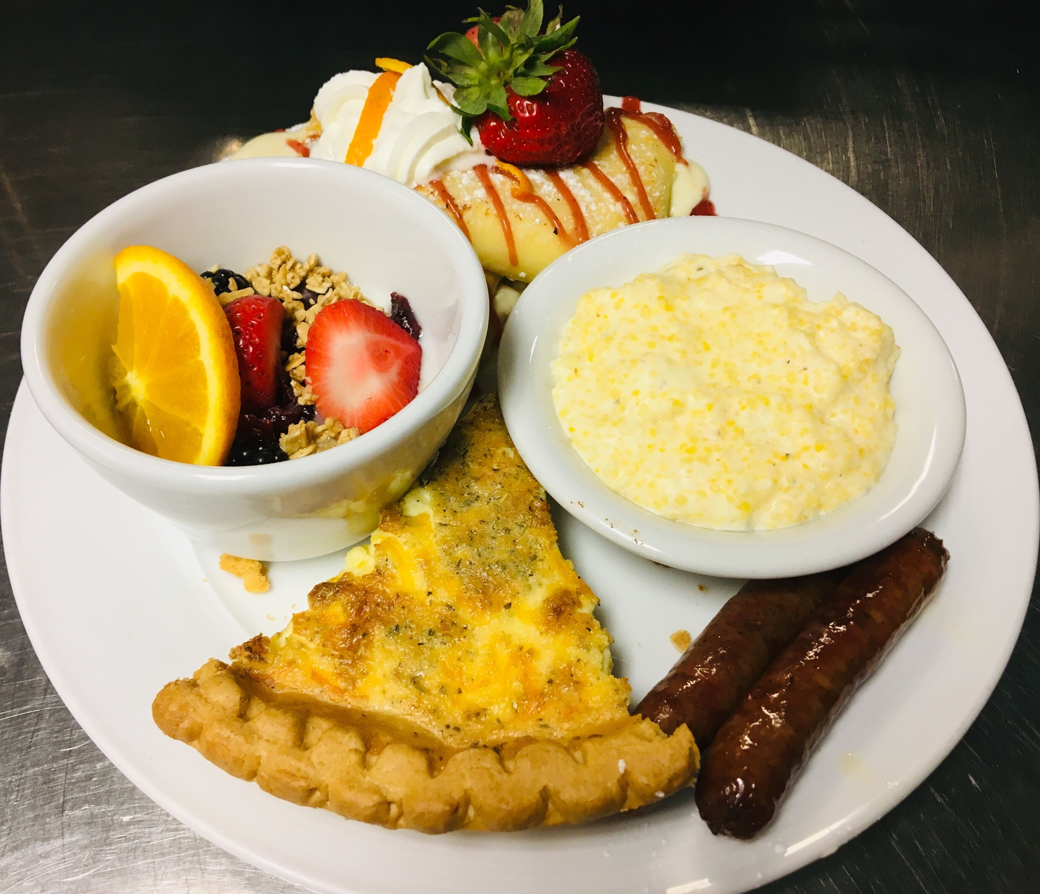 A plate with a slice of quiche, two sausages, a bowl of scrambled eggs, a bowl of fruit salad, and a slice of toast.