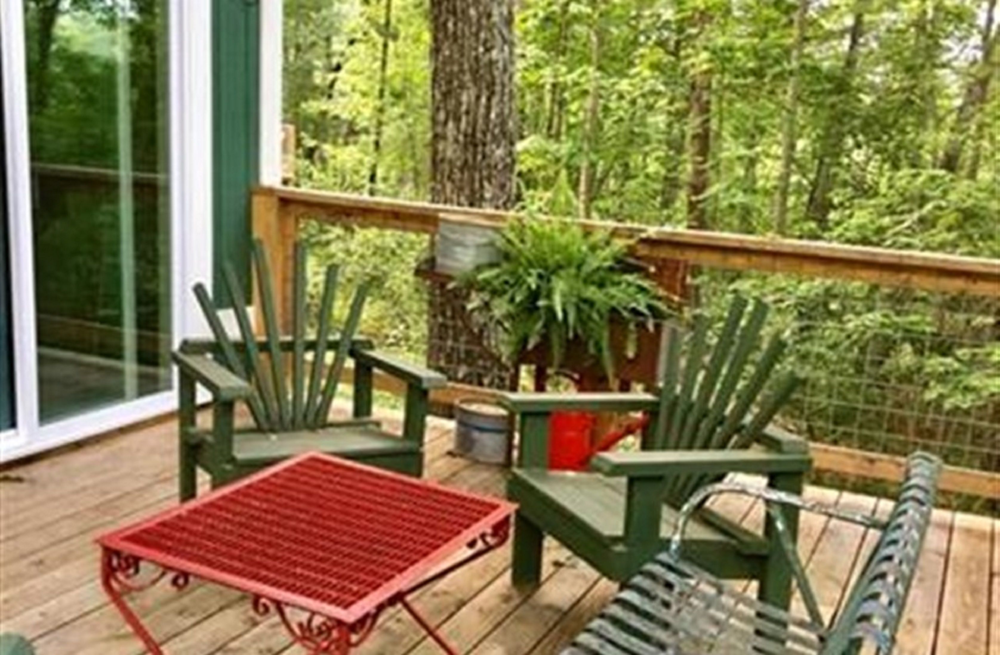 A wooden deck with green chairs and a red table, surrounded by lush greenery.