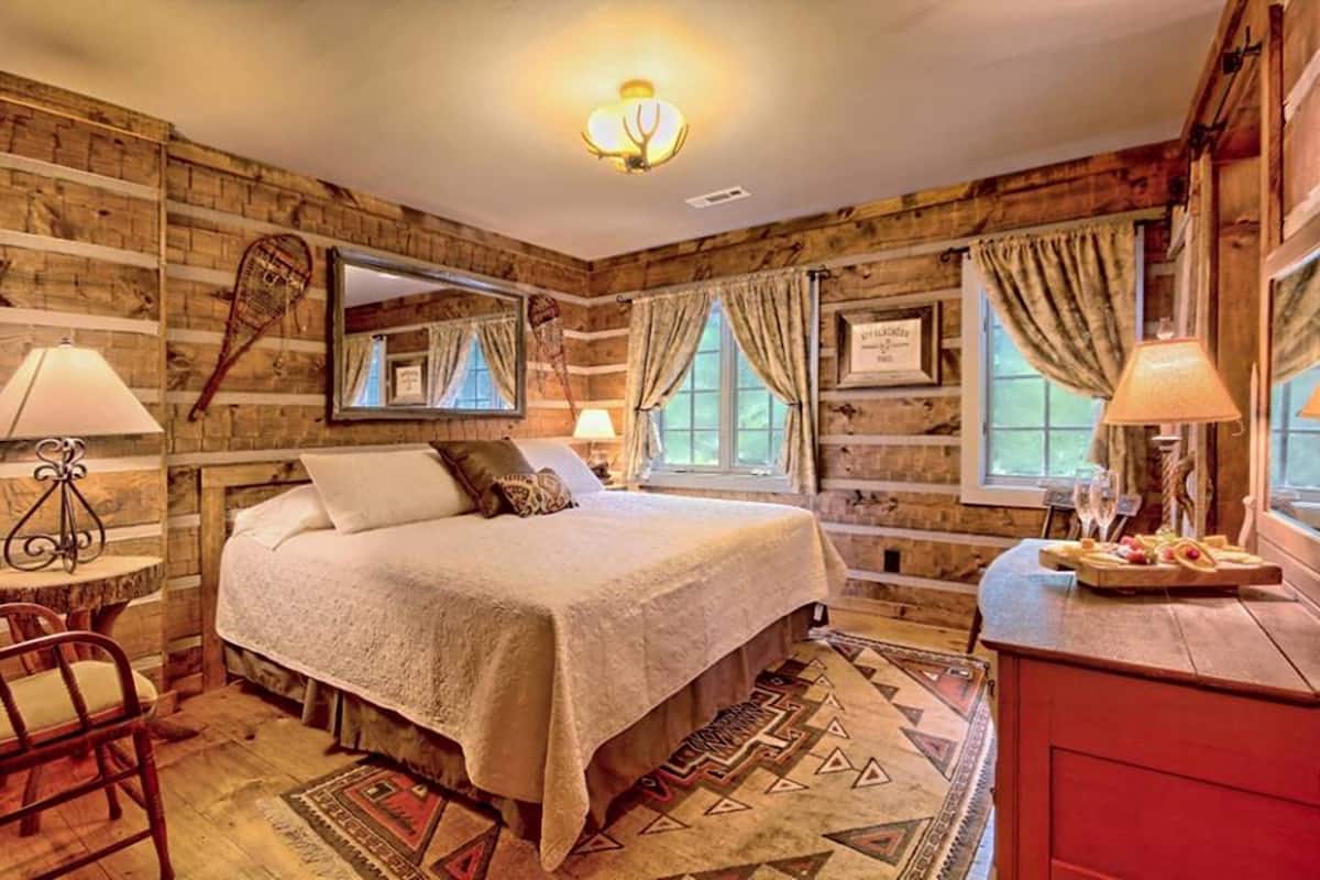 A cozy bedroom with a wooden log wall, a large bed with a white bedspread, two bedside lamps, a wooden dresser, and a window with curtains.