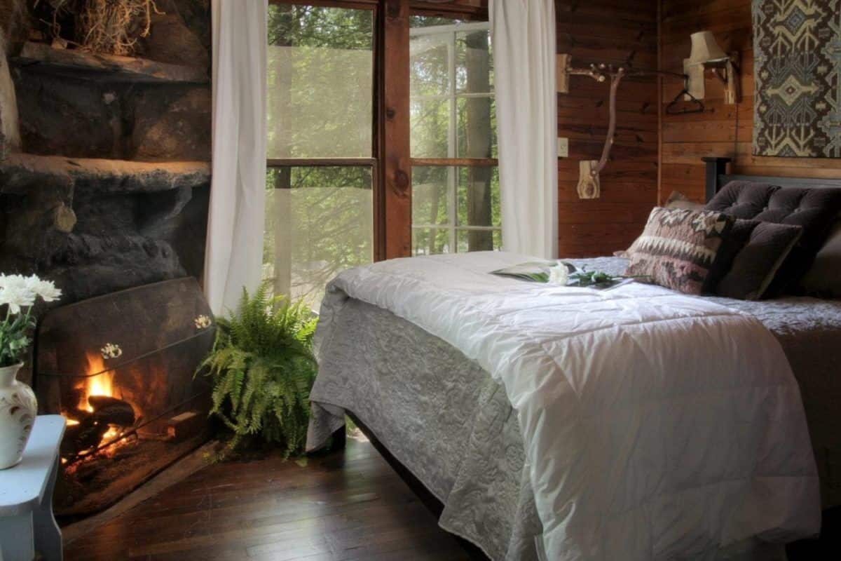A cozy bedroom with a stone fireplace, a large bed with a white bedspread, and a view of the forest through a large window.