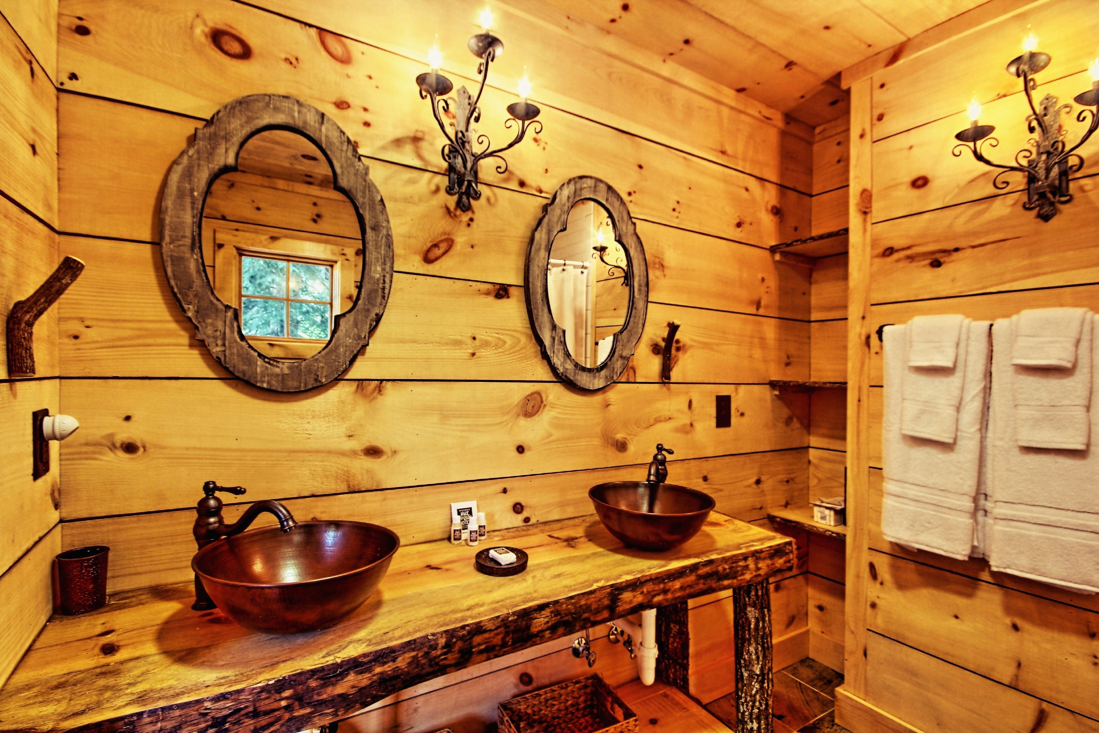 A wooden cabin-style bathroom with two large oval mirrors, a wooden sink with a copper basin, and a towel rack with white towels.