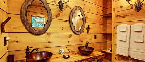 A wooden cabin-style bathroom with two large oval mirrors, a wooden sink with a copper basin, and a towel rack with white towels.