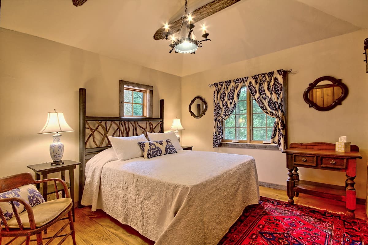 A bedroom with a bed, two bedside tables, a chair, a wooden dresser, a chandelier, a window with curtains, and a mirror on the wall.