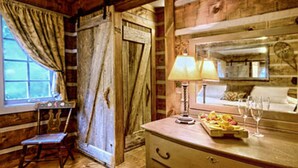 A rustic wooden room with a wooden door, a small table with a lamp, a chair, and a window with curtains.