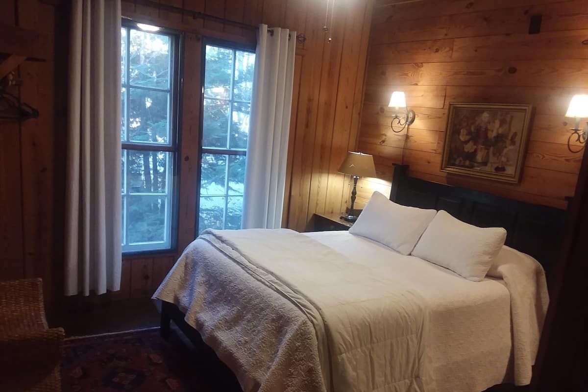 A wooden room with a bed, a nightstand with a lamp, a painting, and a window with curtains.