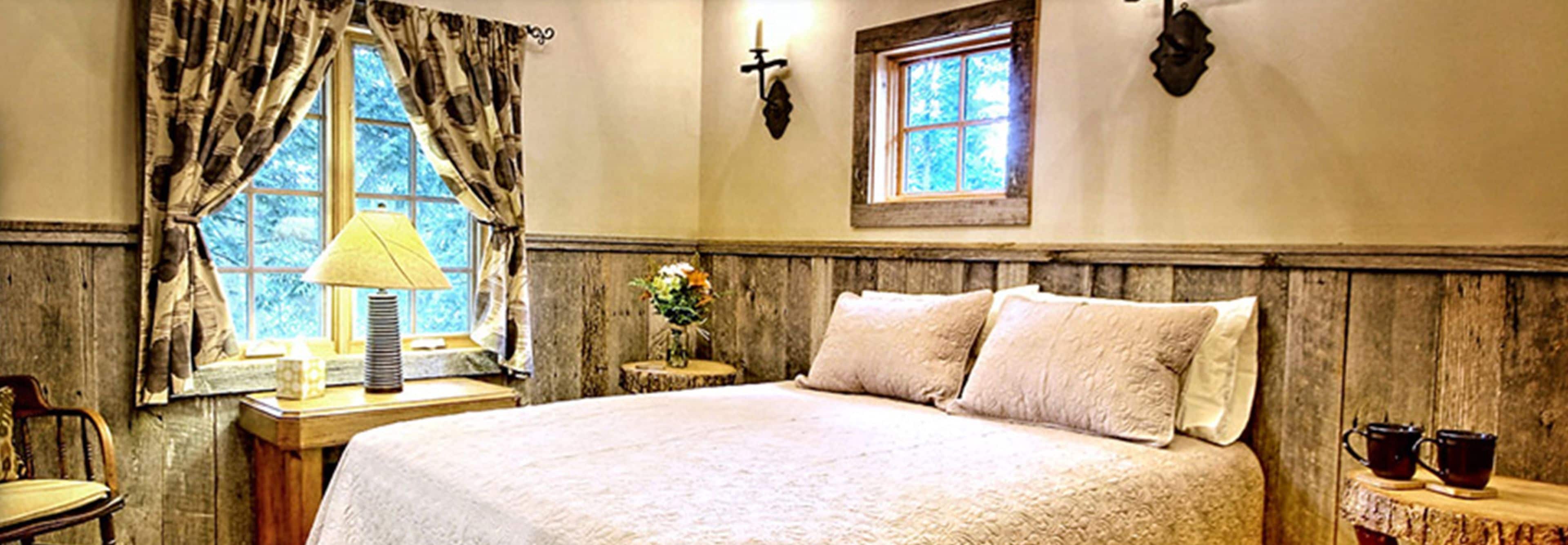 A bedroom with a bed, a nightstand, a chair, a window with curtains, and a wall-mounted light fixture.