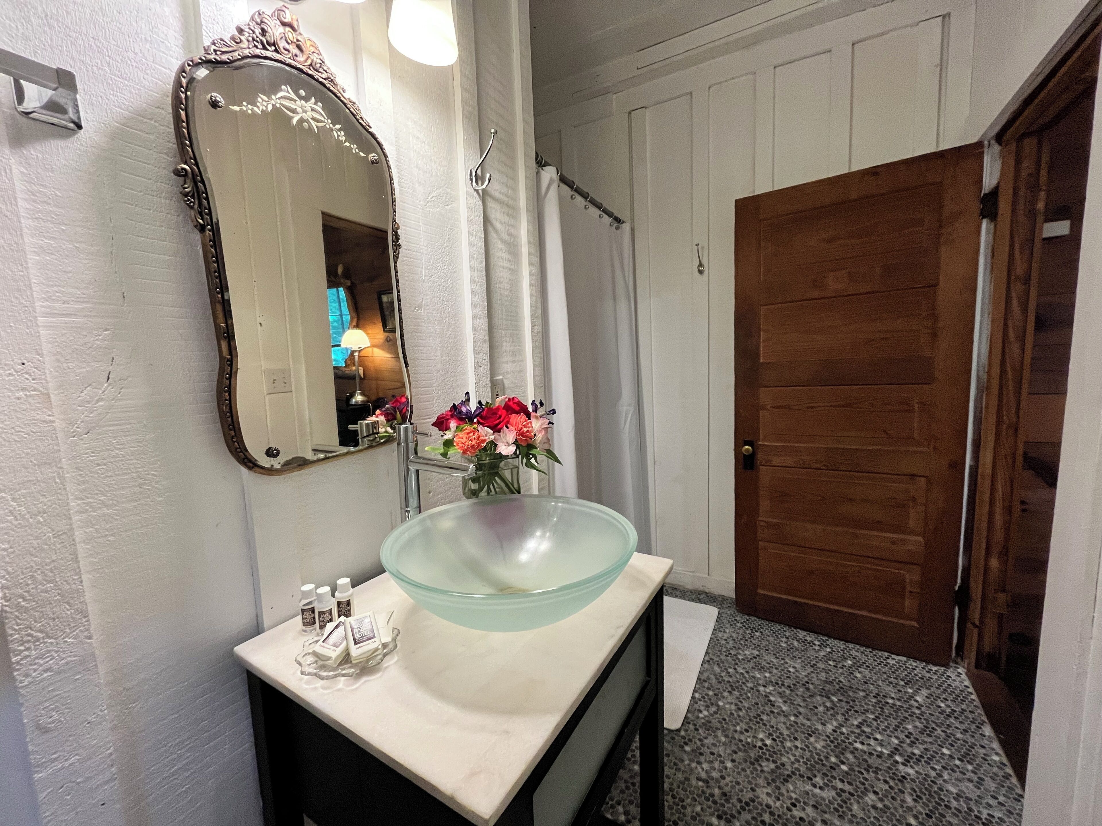 A bathroom with a glass bowl sink, a mirror with ornate frame, and a wooden door.
