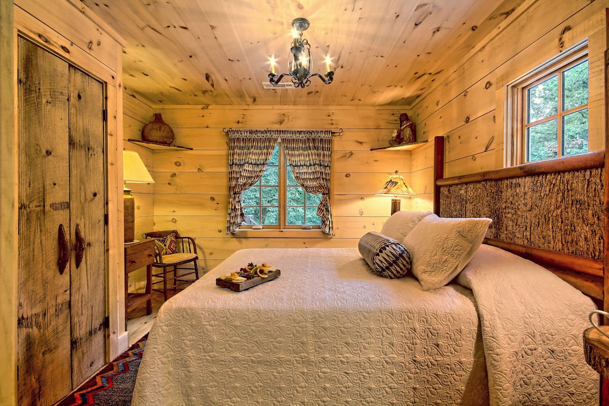 A cozy wooden cabin bedroom with a large bed, a window with curtains, a chandelier, and a small table with a tray of fruit.