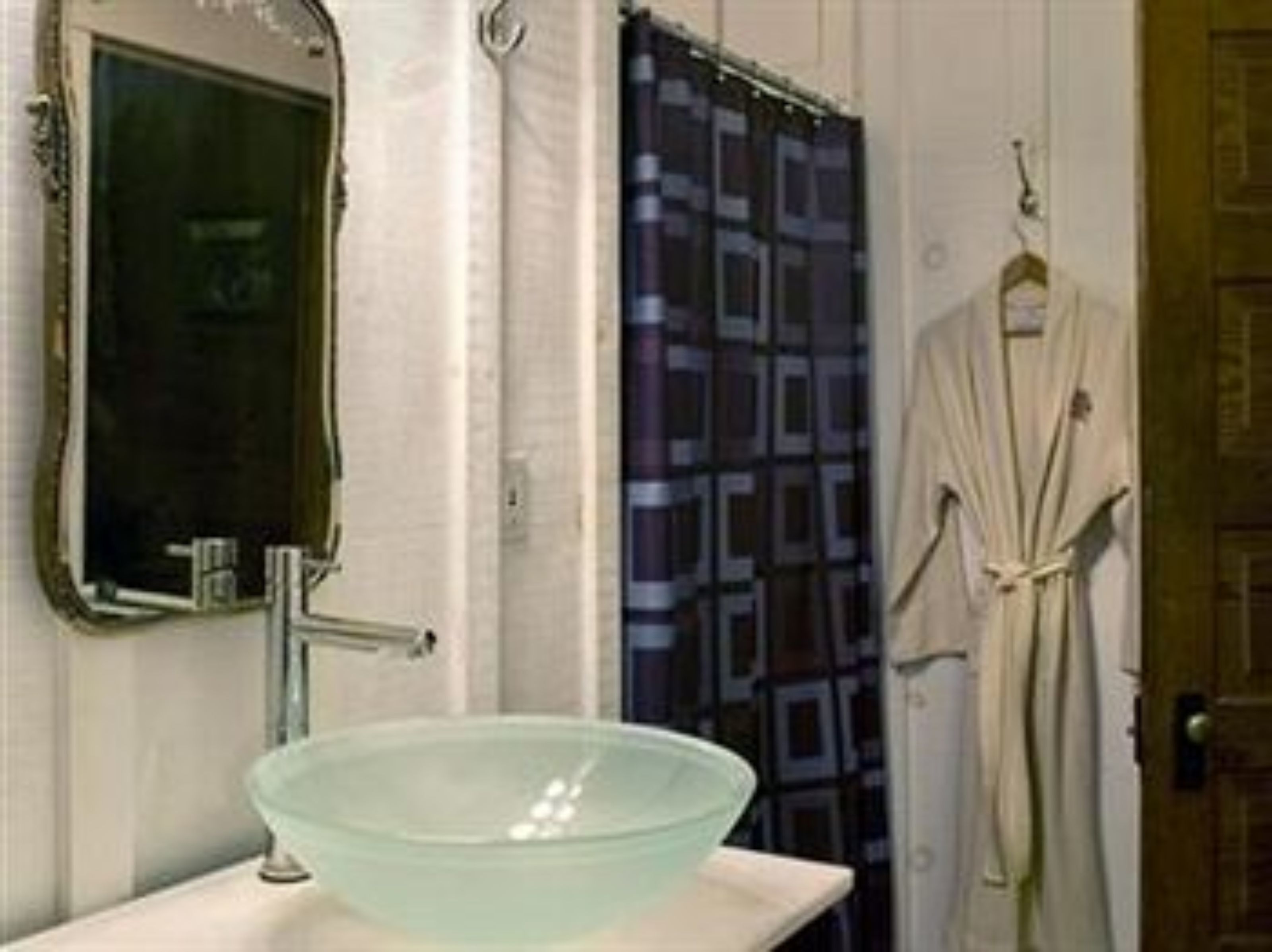 Une salle de bain avec un grand lavabo en forme de bol en verre, un miroir, un rideau de douche et une robe suspendue à une porte.