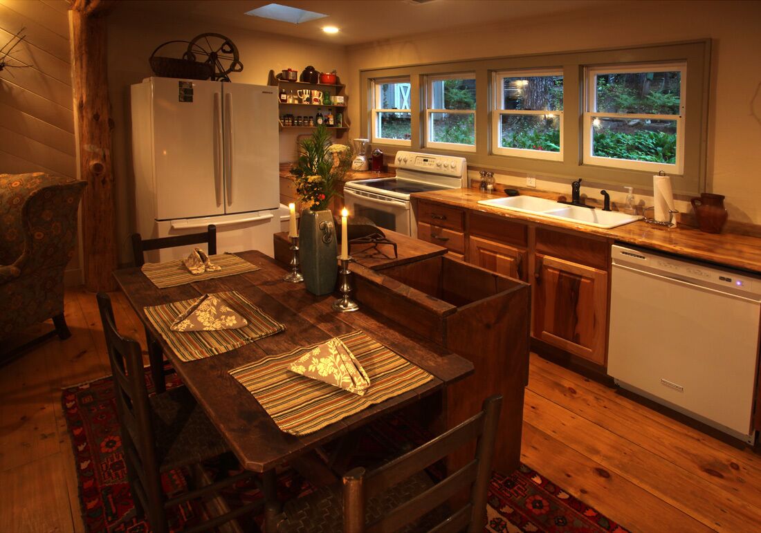 A cozy kitchen with wooden cabinets, a white refrigerator, a stove, and a sink. There are windows with a view of greenery outside.