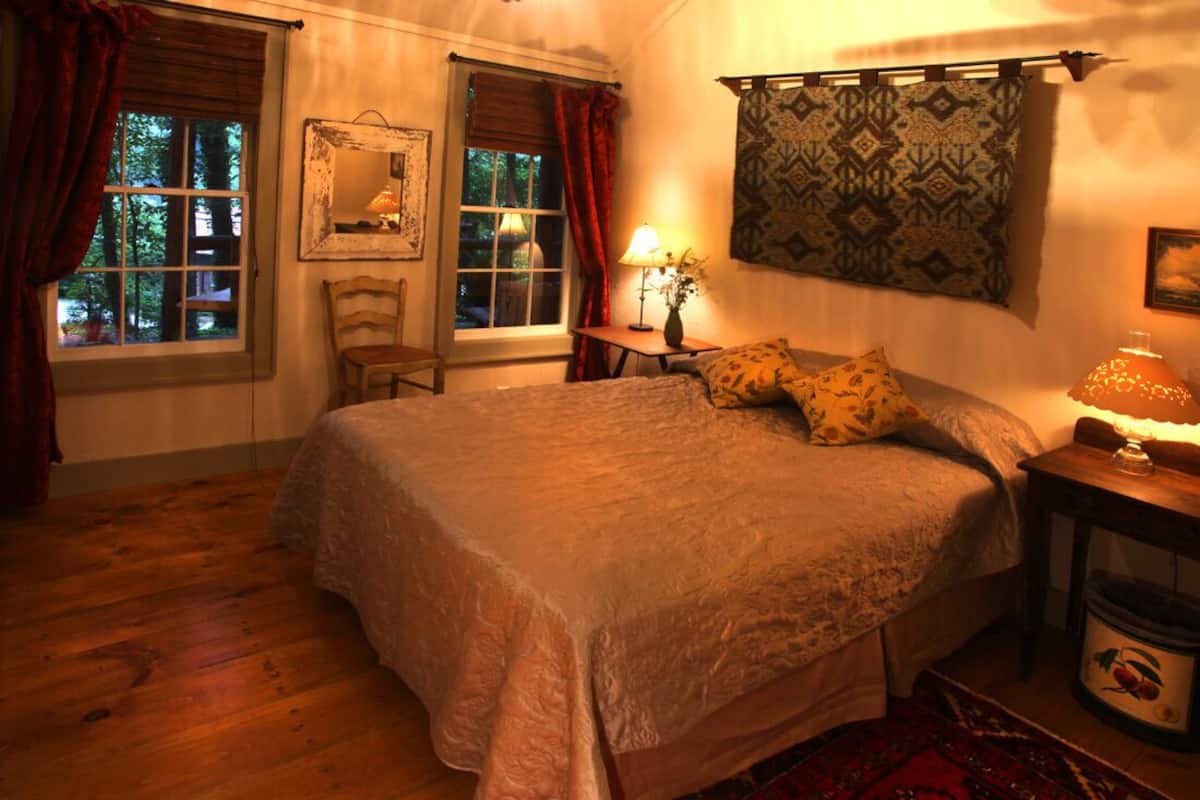 A cozy bedroom with a large bed, a wooden chair, a small table with a lamp, and a patterned curtain.