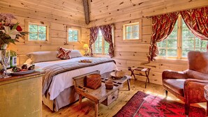 A cozy wooden cabin bedroom with a bed, a wooden table, a chair, and a window with curtains.