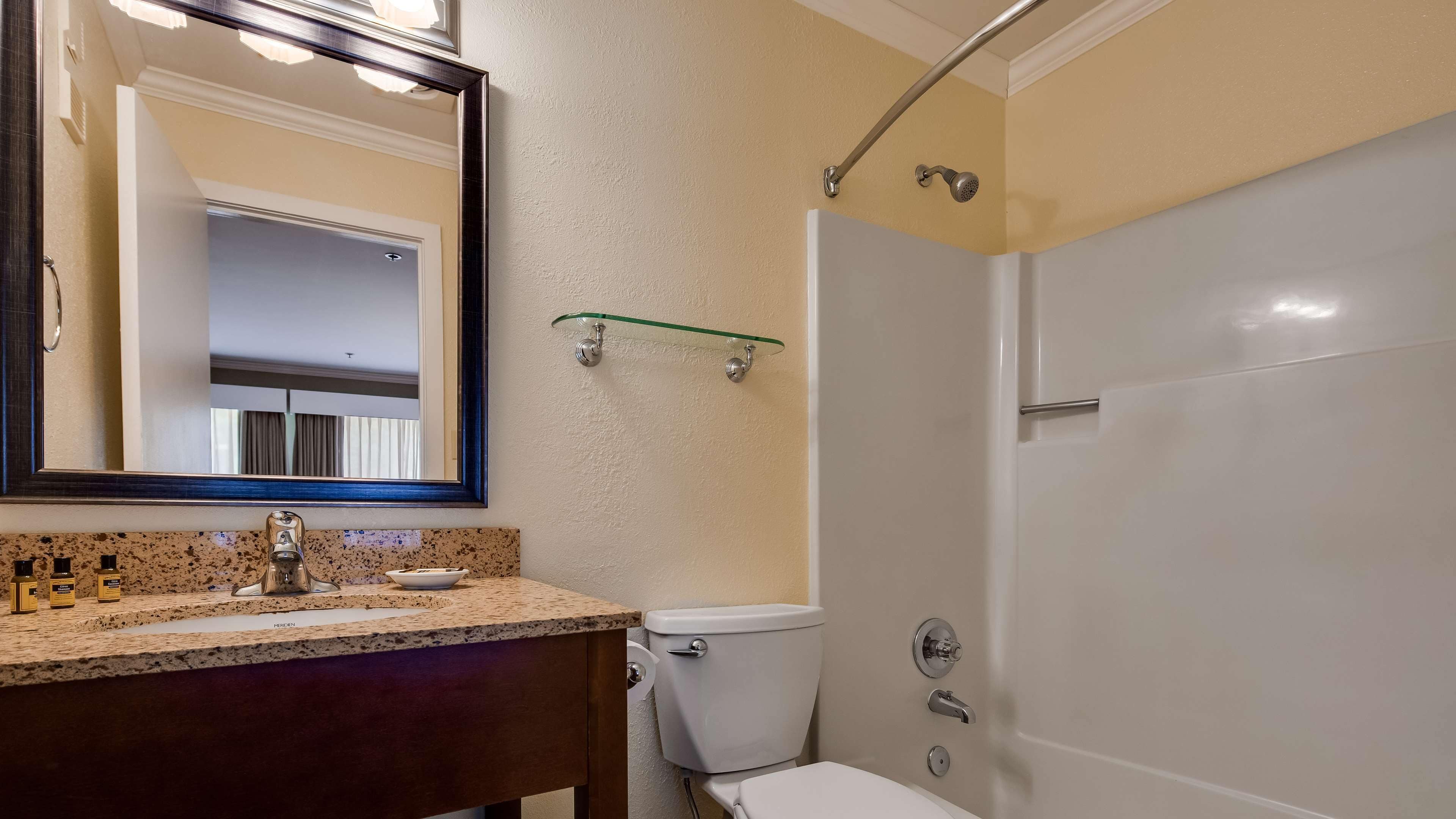 A bathroom with a granite countertop, a mirror, a toilet, a shower, and a glass shelf.