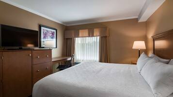 A hotel room with a large bed, a desk with a television, a chair, a lamp, and a window with curtains.