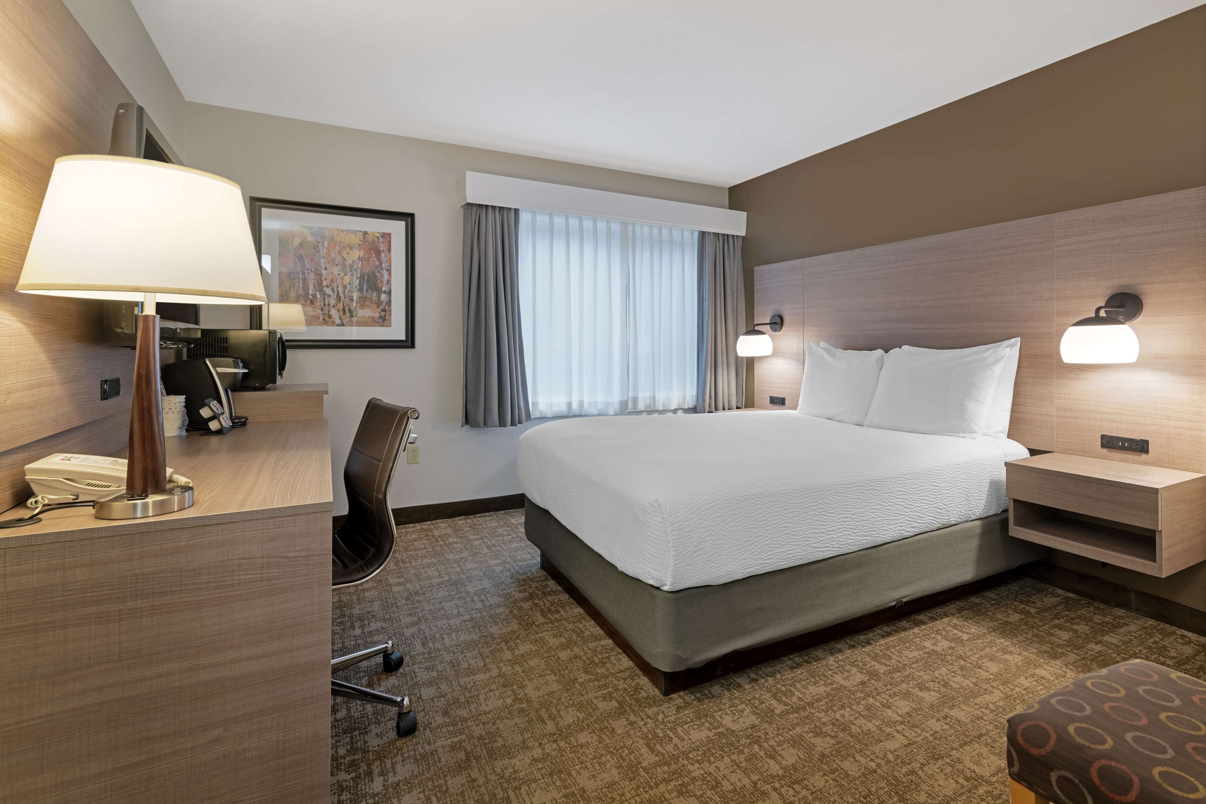 Habitación de hotel con una cama grande, un escritorio con lámpara, una silla, una lámpara de escritorio, una mesita de noche con lámpara y una ventana con cortinas.