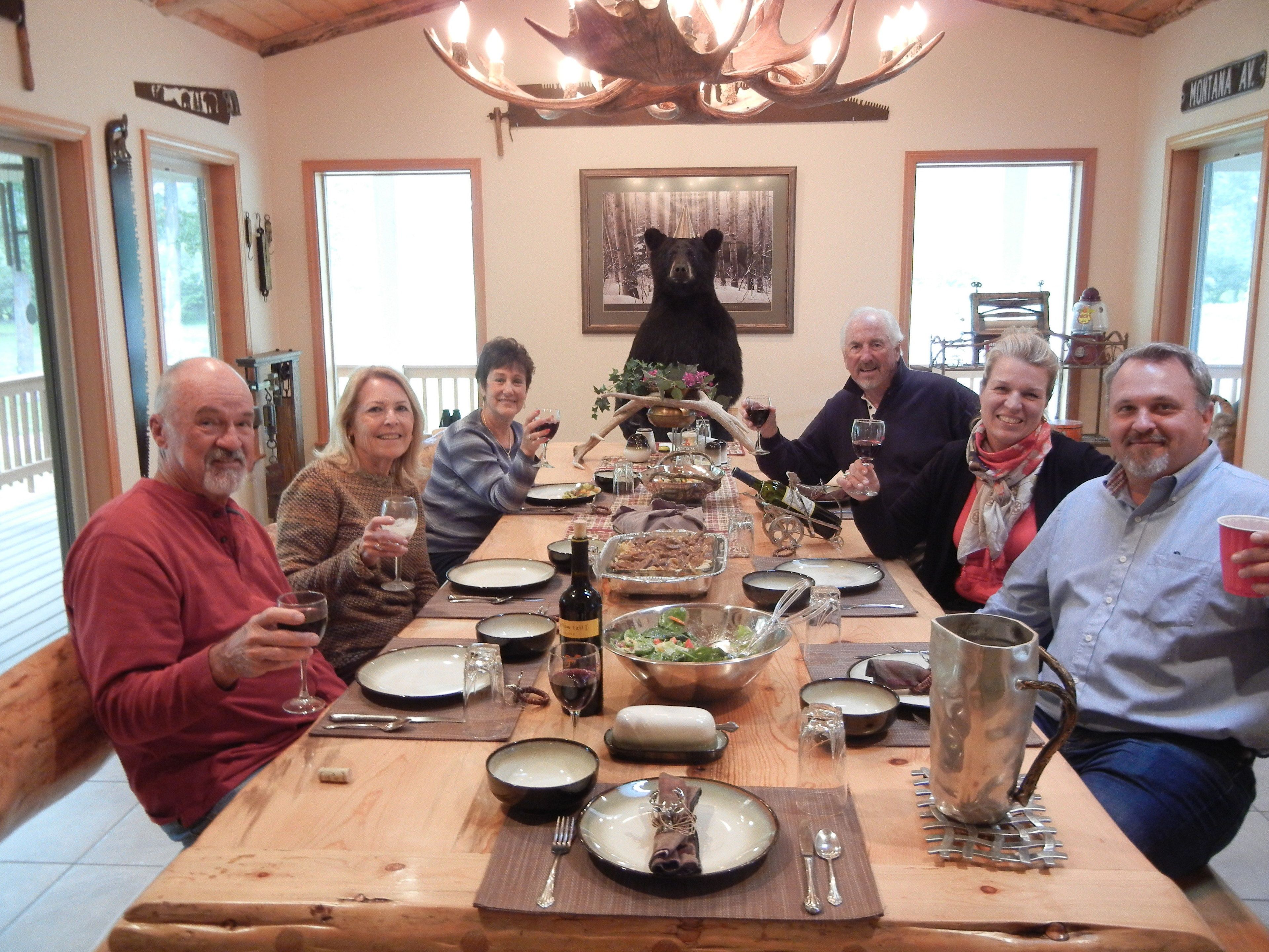 A group of people seated around a wooden dining table with various dishes, glasses, and a bottle of wine.