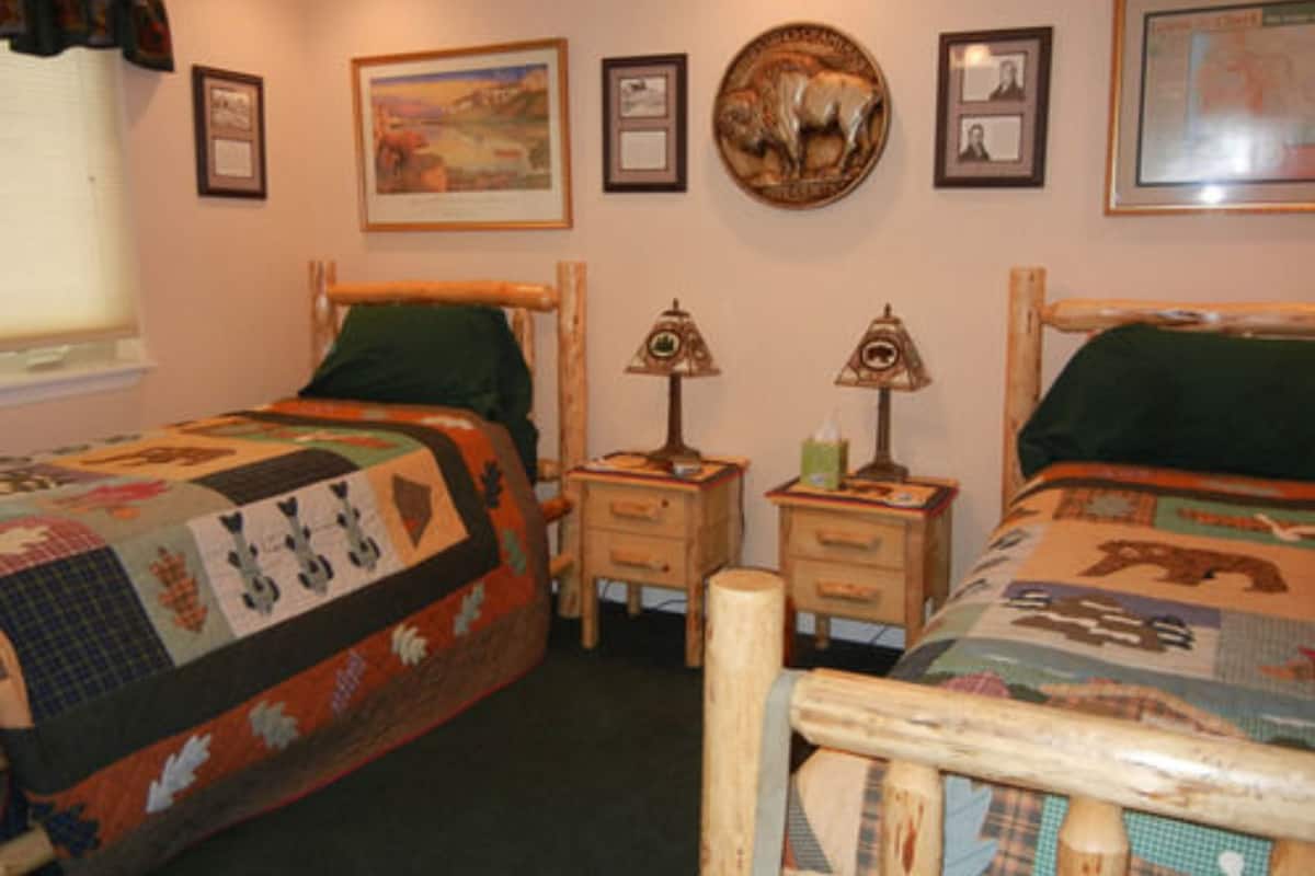 Una habitación con dos camas de madera, cada una con una colcha verde y un edredón. Hay cuadros enmarcados en la pared, una mesita de noche de madera con una lámpara y una ventana con cortinas.