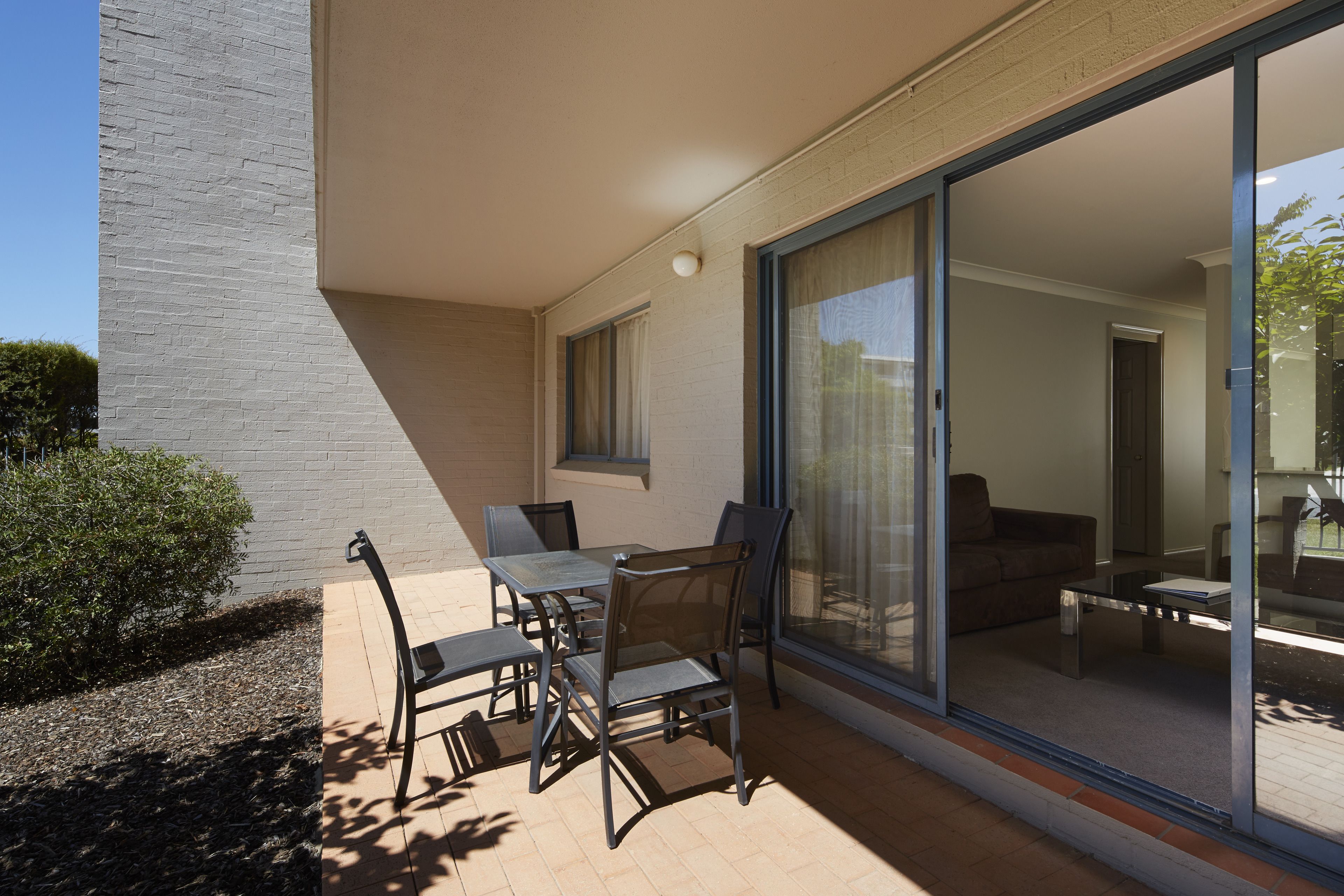 Een buitenpatio met een tafel en stoelen, een schuifpui die leidt naar een woonruimte en een muur van witte bakstenen.