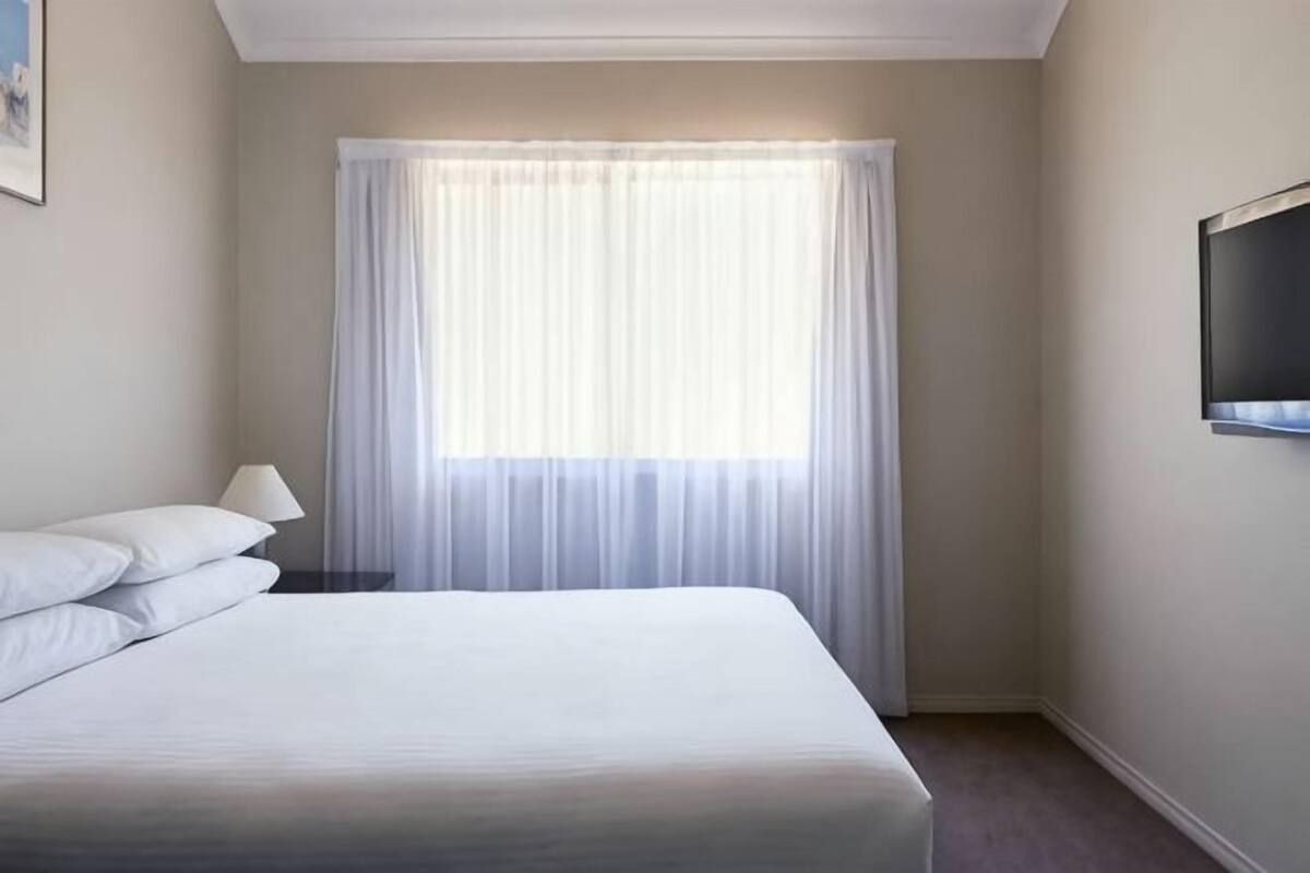 Una habitación de hotel con una cama, una ventana con cortinas translúcidas, un televisor de pantalla plana montado en la pared y una mesita de noche con una lámpara.