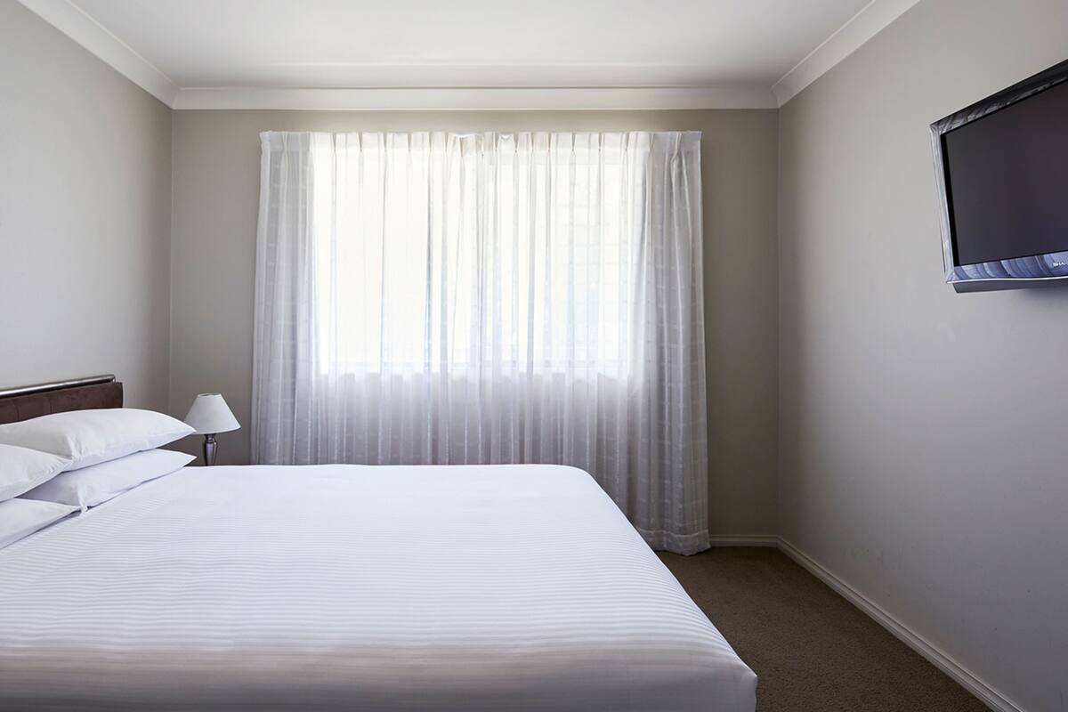 Habitación de hotel con una cama grande, un televisor de pantalla plana en la pared y una ventana con cortinas translúcidas.