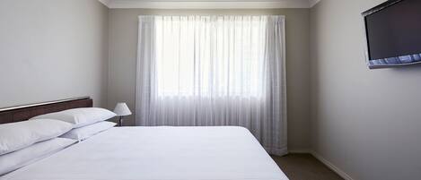 A hotel room with a large bed, a flat-screen TV mounted on the wall, and a window with sheer curtains.