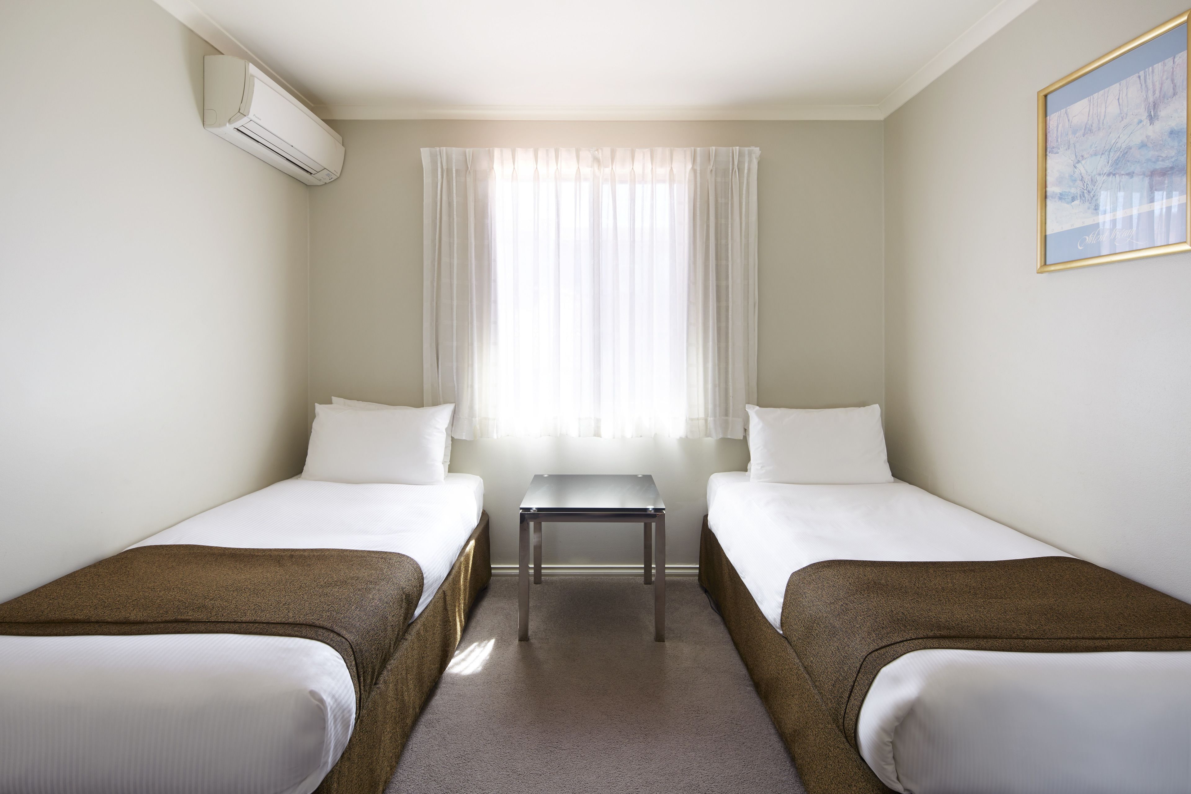 A hotel room with two single beds, a small table, and a window with sheer curtains.