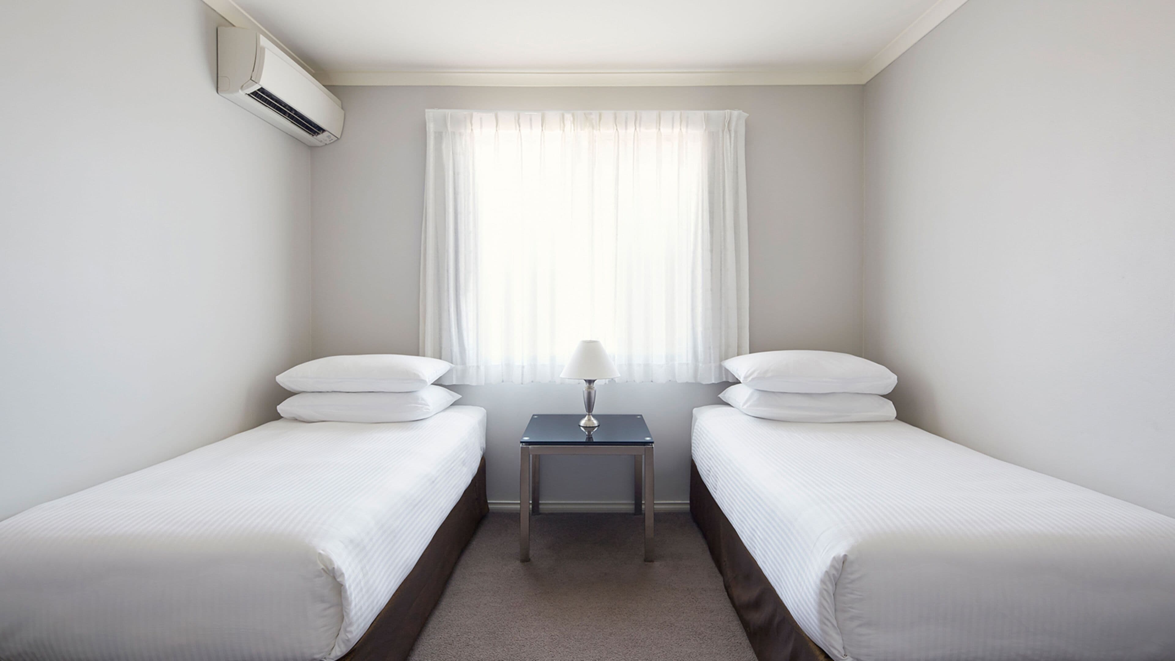 A hotel room with two single beds, a small table with a lamp, and an air conditioning unit on the wall.