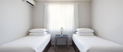 A hotel room with two single beds, a small table with a lamp, and an air conditioning unit on the wall.