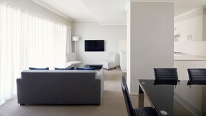 Una habitación de hotel moderna con un sofá gris, una pequeña mesa de centro, un televisor montado en la pared y una zona de comedor con una mesa de cristal y sillas negras.