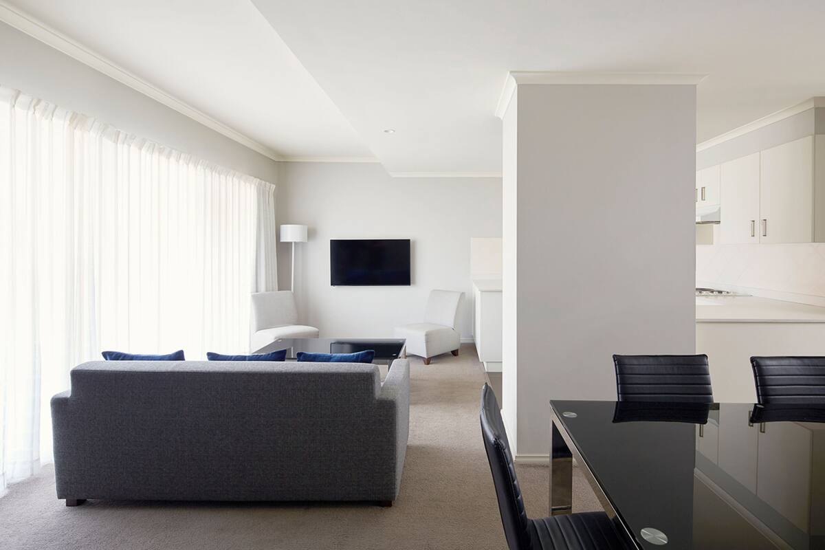 Una habitación de hotel moderna con un sofá gris, una pequeña mesa de centro, un televisor montado en la pared y una zona de comedor con una mesa de cristal y sillas negras.