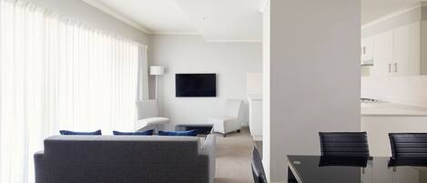 A modern hotel room with a grey sofa, a small coffee table, a TV mounted on the wall, and a dining area with a glass table and black chairs.