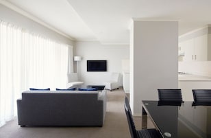 Quarto de hotel moderno com sofá cinzento, uma pequena mesa de centro, televisão presa na parede e um espaço de refeições com mesa de vidro e cadeiras pretas.