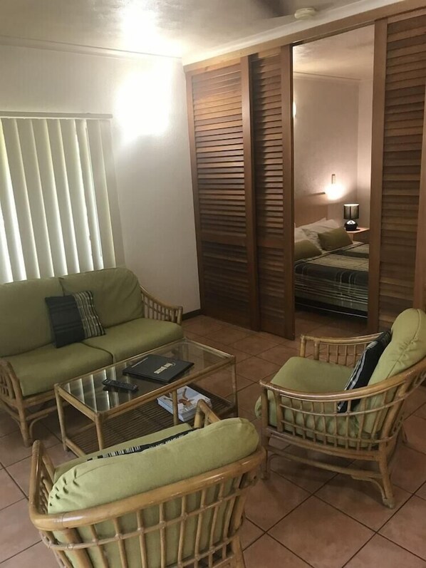 A room with a sofa, two wicker chairs, a glass coffee table, and a bed with a wooden headboard. - Palm Villas Resort (Port Douglas)