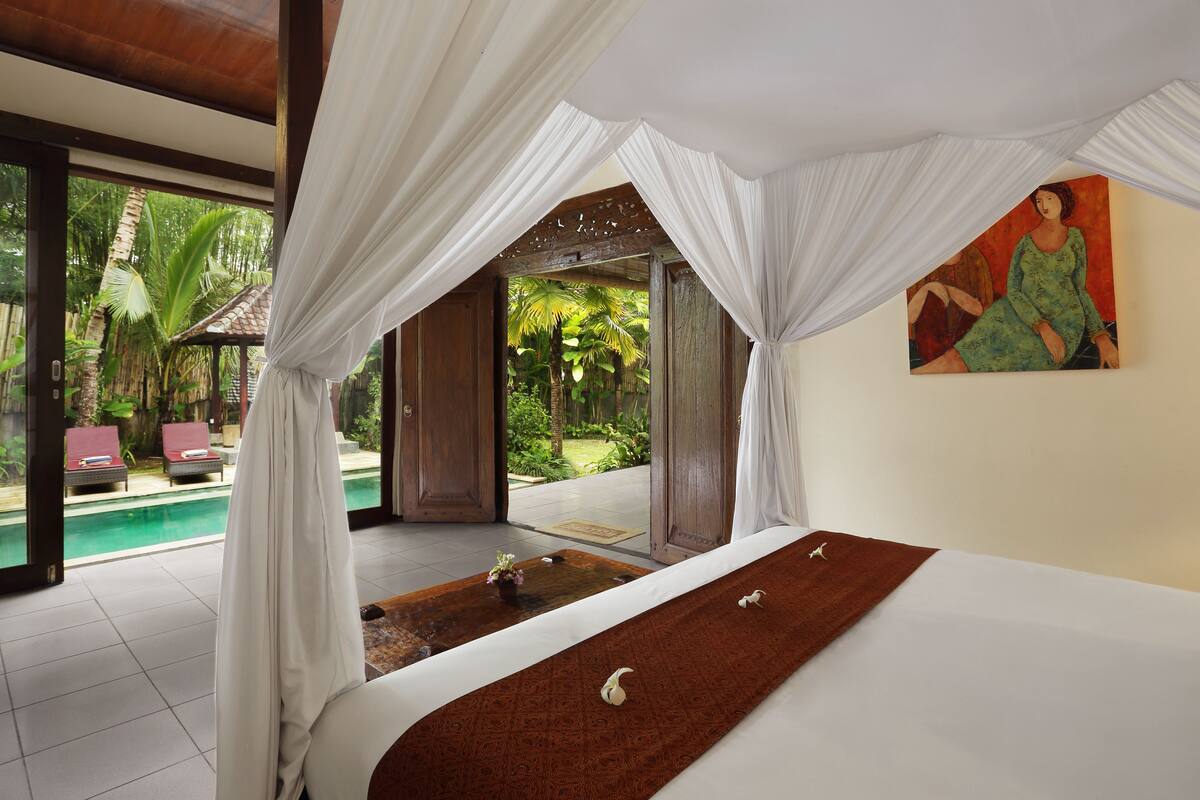 A bedroom with a bed, white curtains, a painting, and a view of a pool and garden.