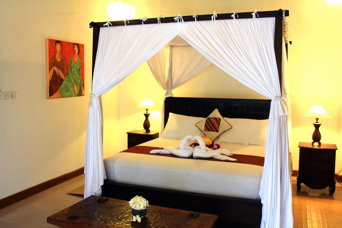 A four-poster canopy bed with white linens, two bedside lamps, a decorative pillow, and a small table with a flower arrangement.