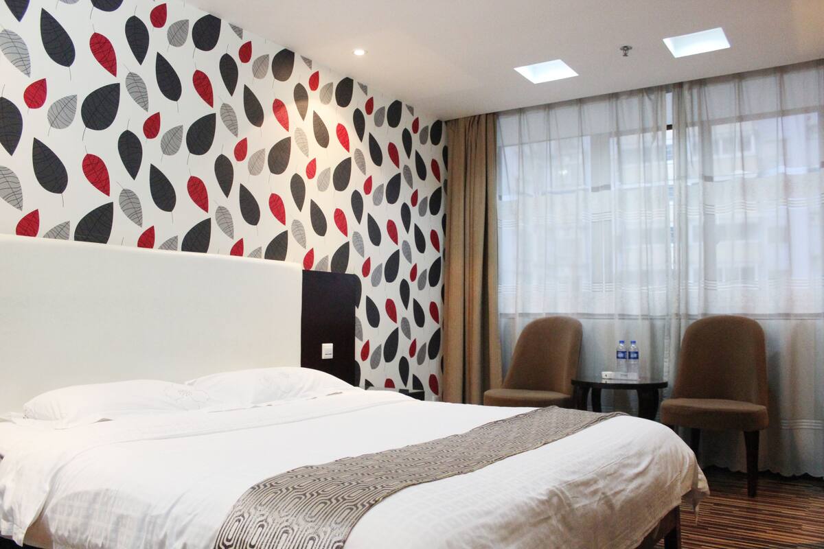 A hotel room with a large bed, two chairs, a small table with water bottles, and a patterned wall.