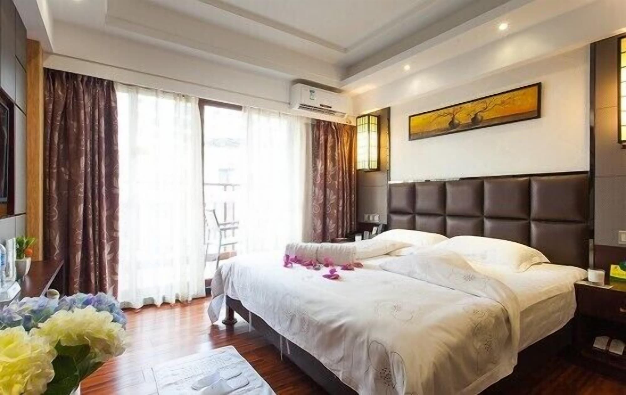 A hotel room with a large bed, a headboard, an air conditioning unit, a painting on the wall, and a window with curtains.
