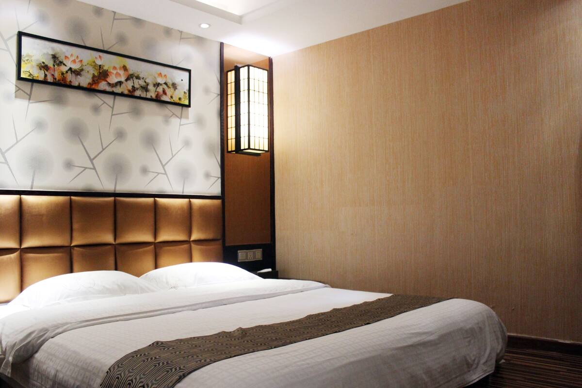 A hotel room with a large bed, a headboard with a geometric pattern, a wall-mounted artwork, and a window with a view of the outside.