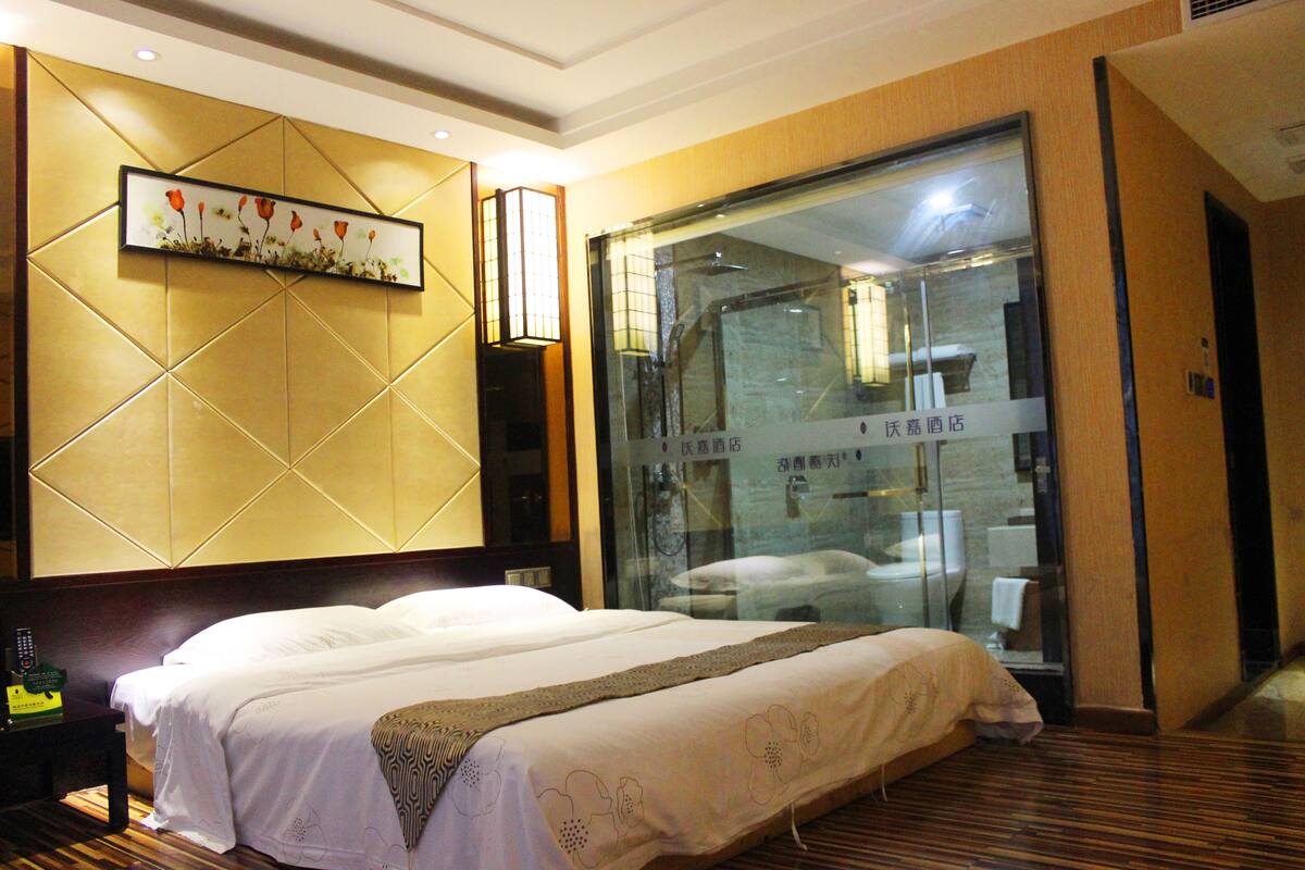 A hotel room with a large bed, a wall-mounted artwork, and a glass partition with a bathroom visible.