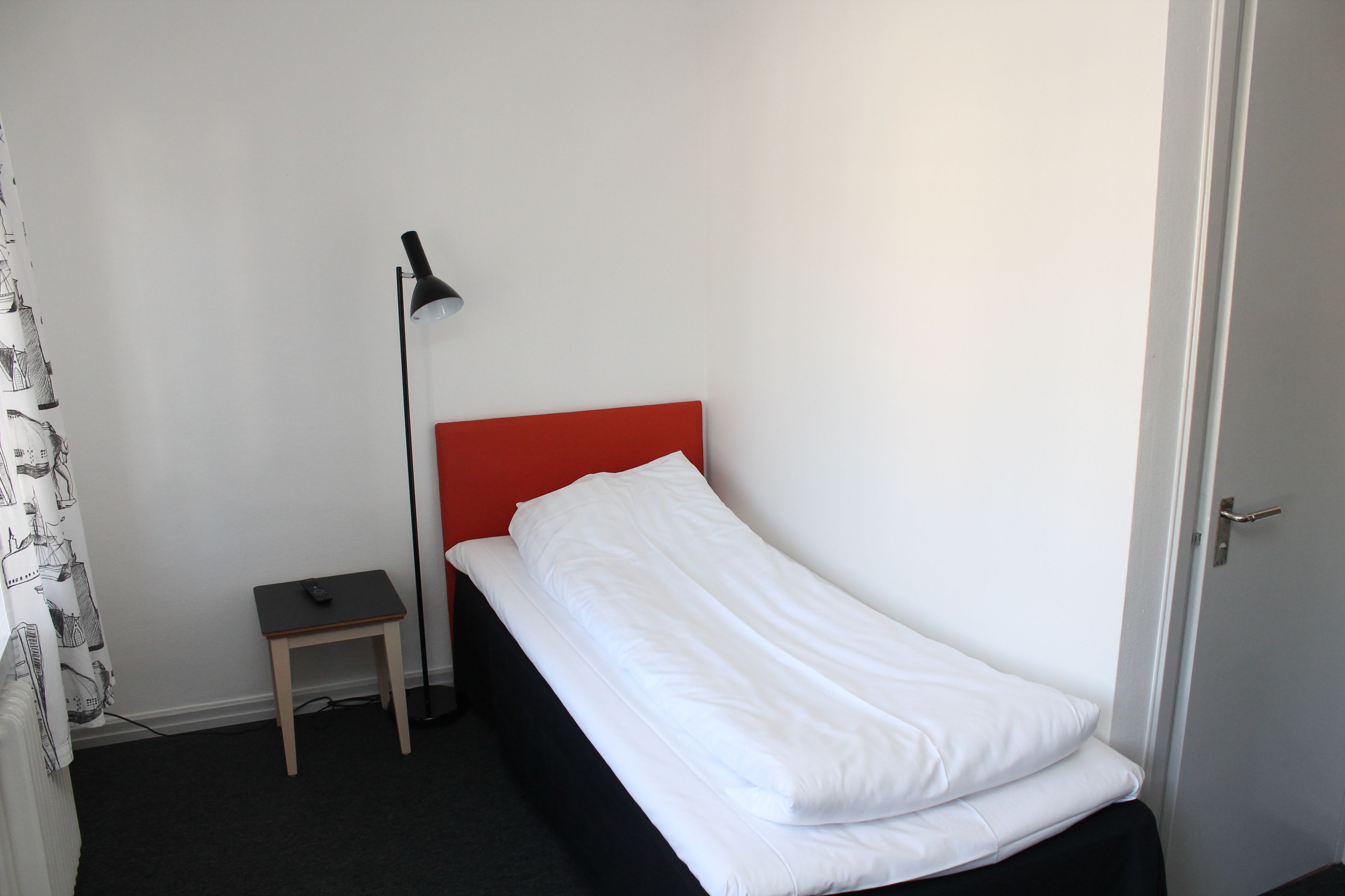 A small, single-bed room with a red headboard, a bedside table, a lamp, and a door.
