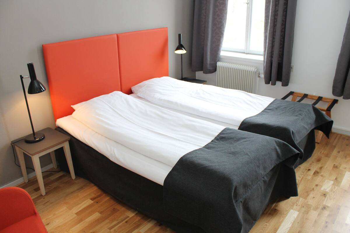 A double bed with a red headboard, a bedside table with a lamp, and a window with curtains.