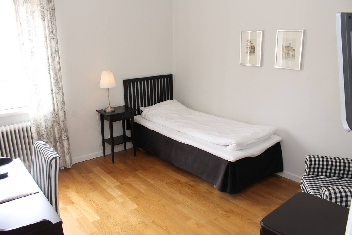 A small, neatly arranged bedroom with a single bed, a bedside table with a lamp, a chair, and a radiator.