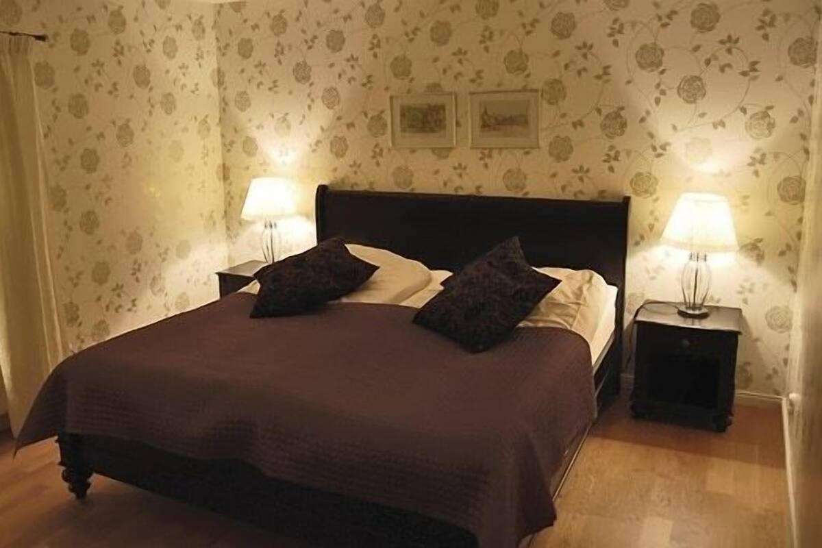 A neatly made bed with a dark brown bedspread and two pillows, flanked by two bedside tables with lamps, in a room with floral wallpaper and a window with curtains.