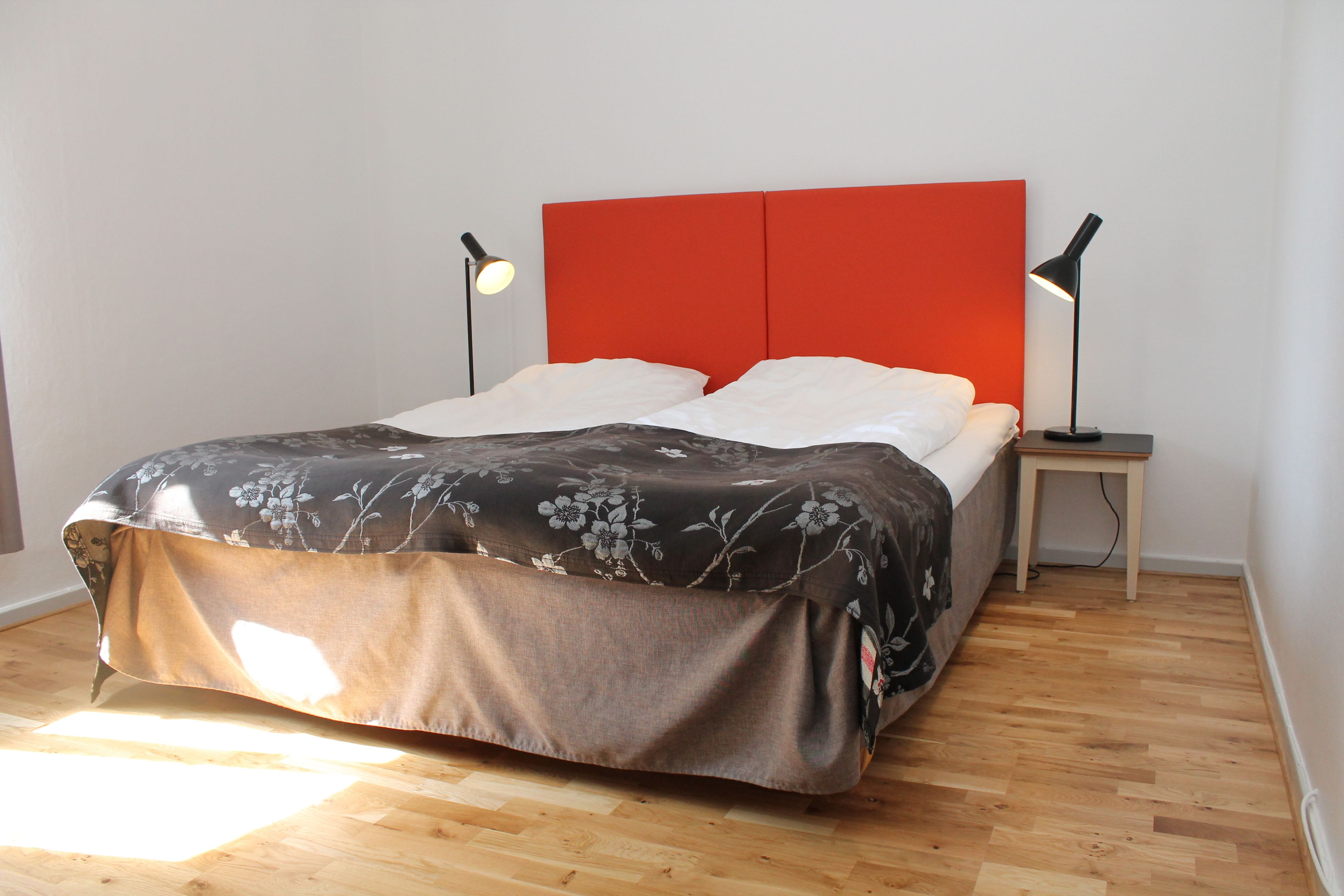 Una cama con colcha estampada, dos lámparas de noche y un cabecero rojo en una habitación con suelo de madera.