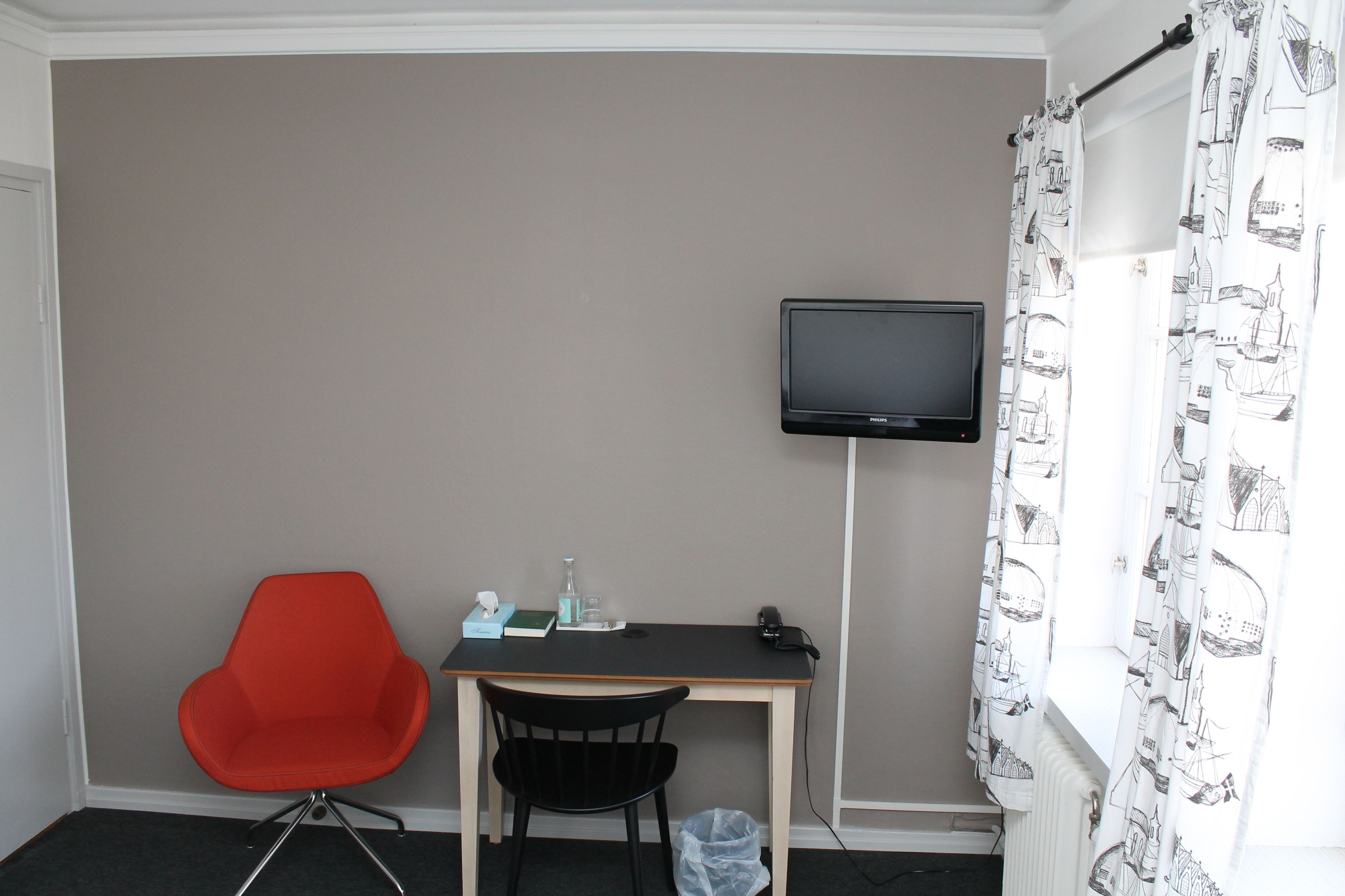 Une petite pièce comprenant un bureau, une chaise rouge, une chaise noire, un téléviseur fixé au mur et une fenêtre ornée de rideaux à motifs.