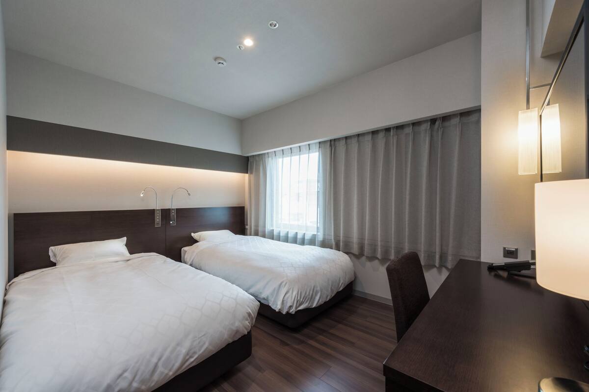 Hotelkamer met twee bedden, een bureau, een stoel, een lamp en een raam met gordijnen.