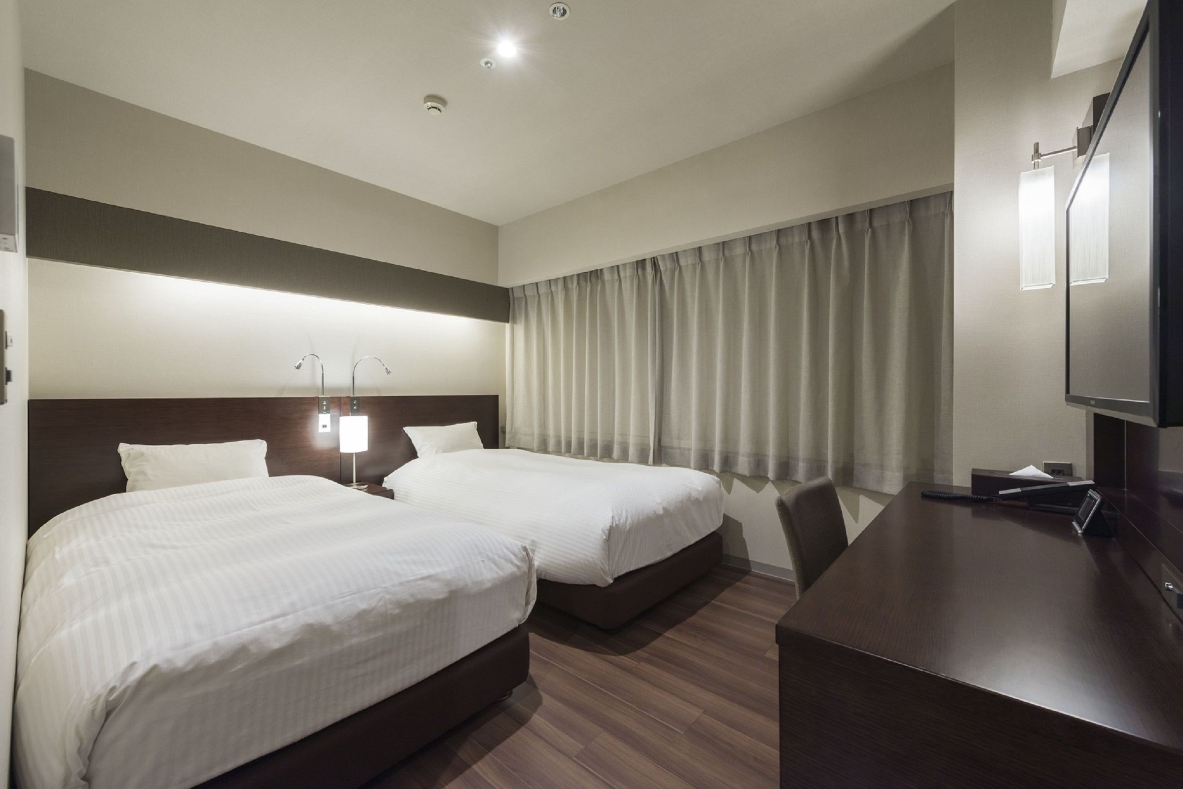 Ein Hotelzimmer mit zwei Betten, einem Schreibtisch, einem Fernseher und einem Fenster mit Vorhängen.