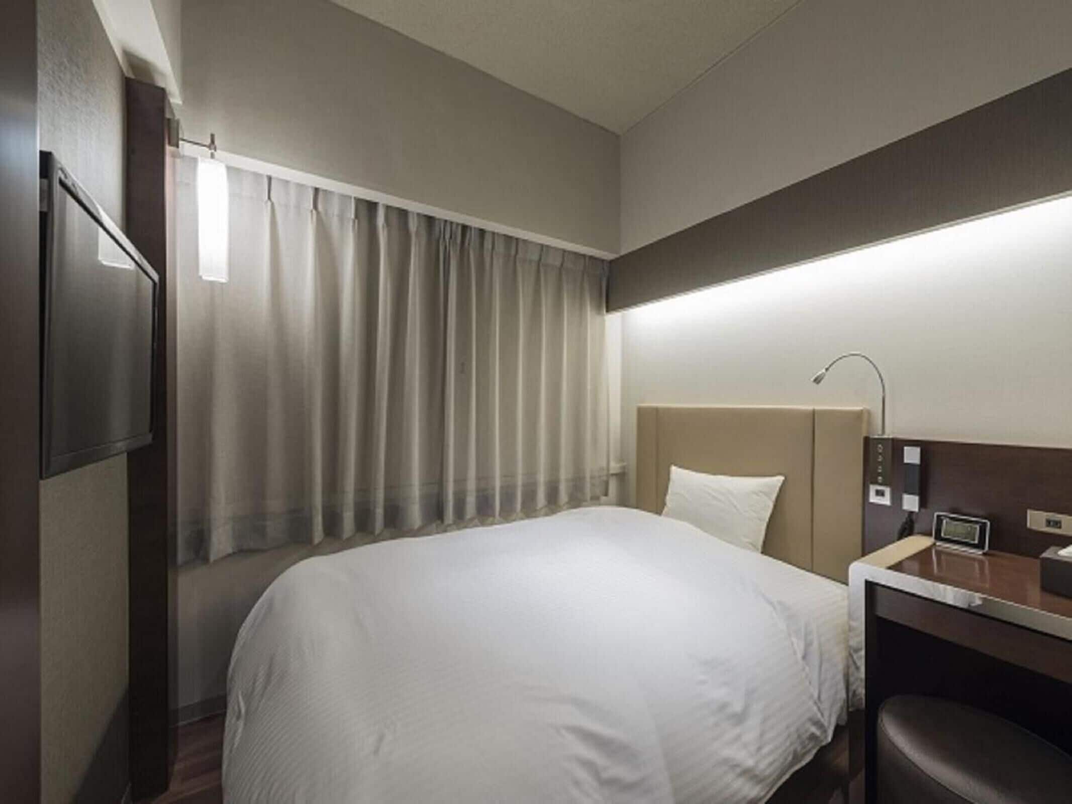 A hotel room with a bed, a desk, a chair, a TV, and a window with curtains.