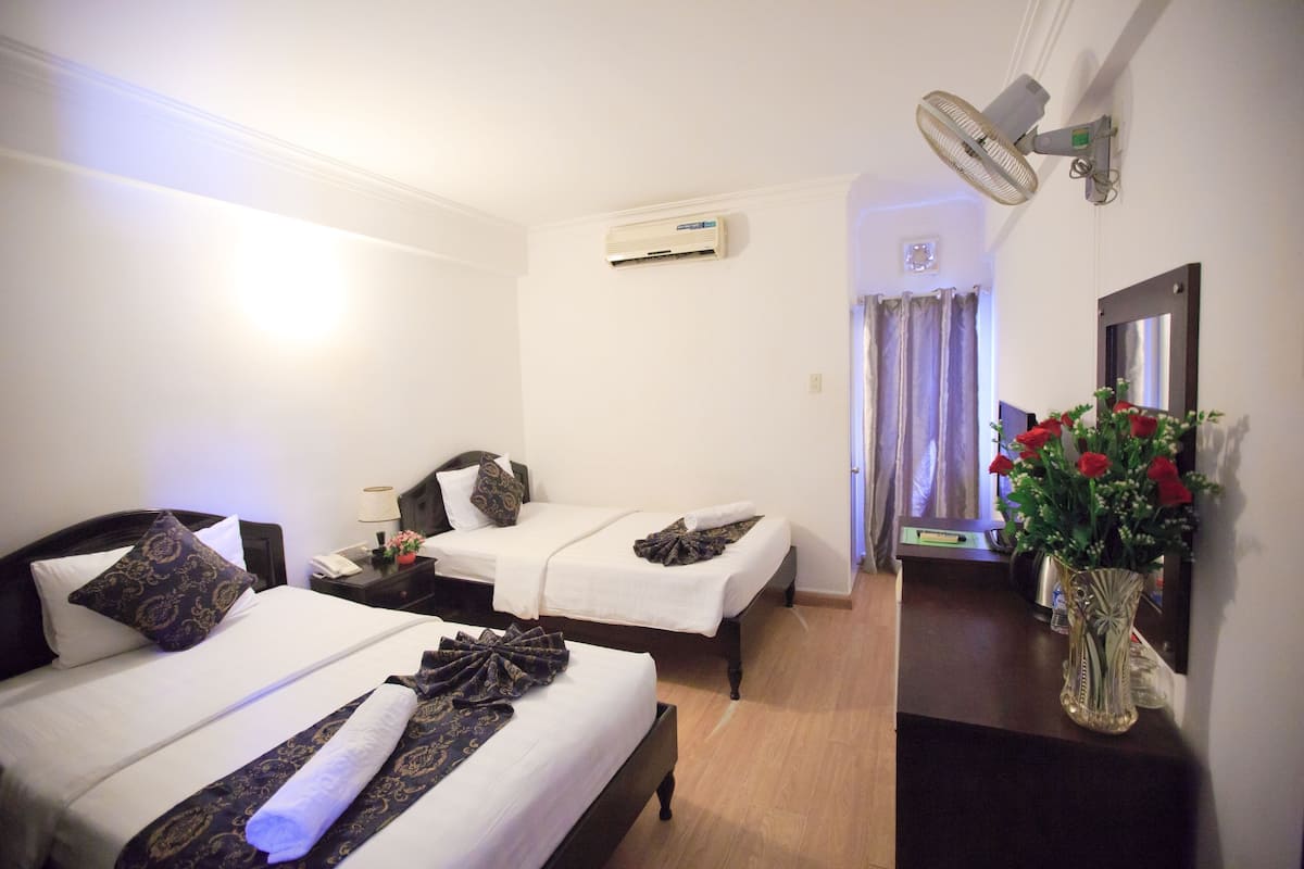 Une chambre d’hôtel avec deux lits, une table de chevet avec une lampe, un climatiseur, un ventilateur fixé au mur, un miroir et un vase avec des fleurs.